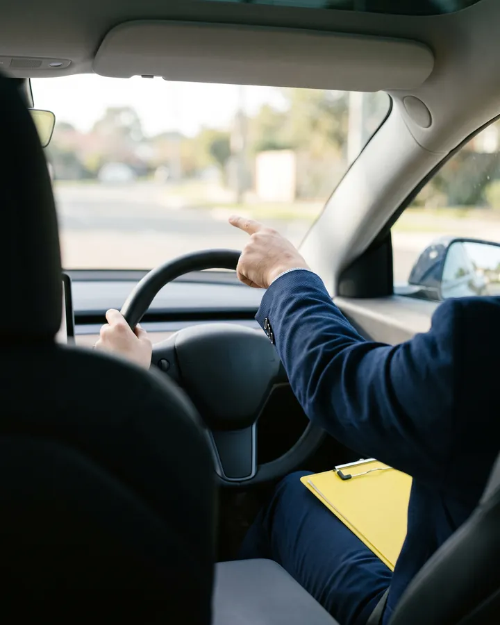 Fahrlehrer zeigt im Auto nach vorn, auf dem Schoß ein gelbes Klemmbrett