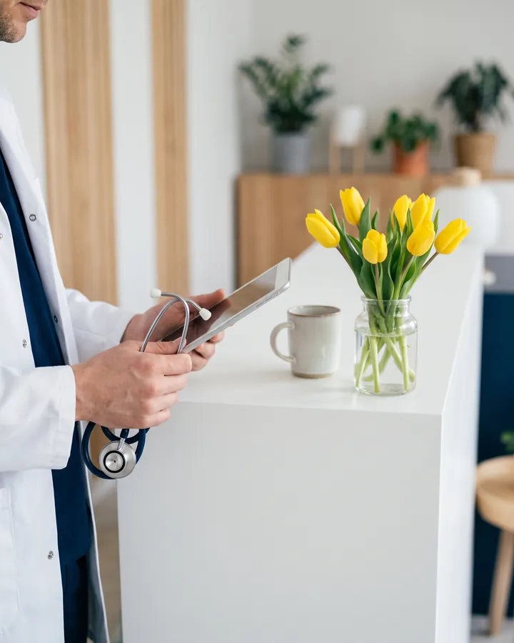 Arzt mit Stethoskop und Tablet am Empfang, daneben gelbe Tulpen
